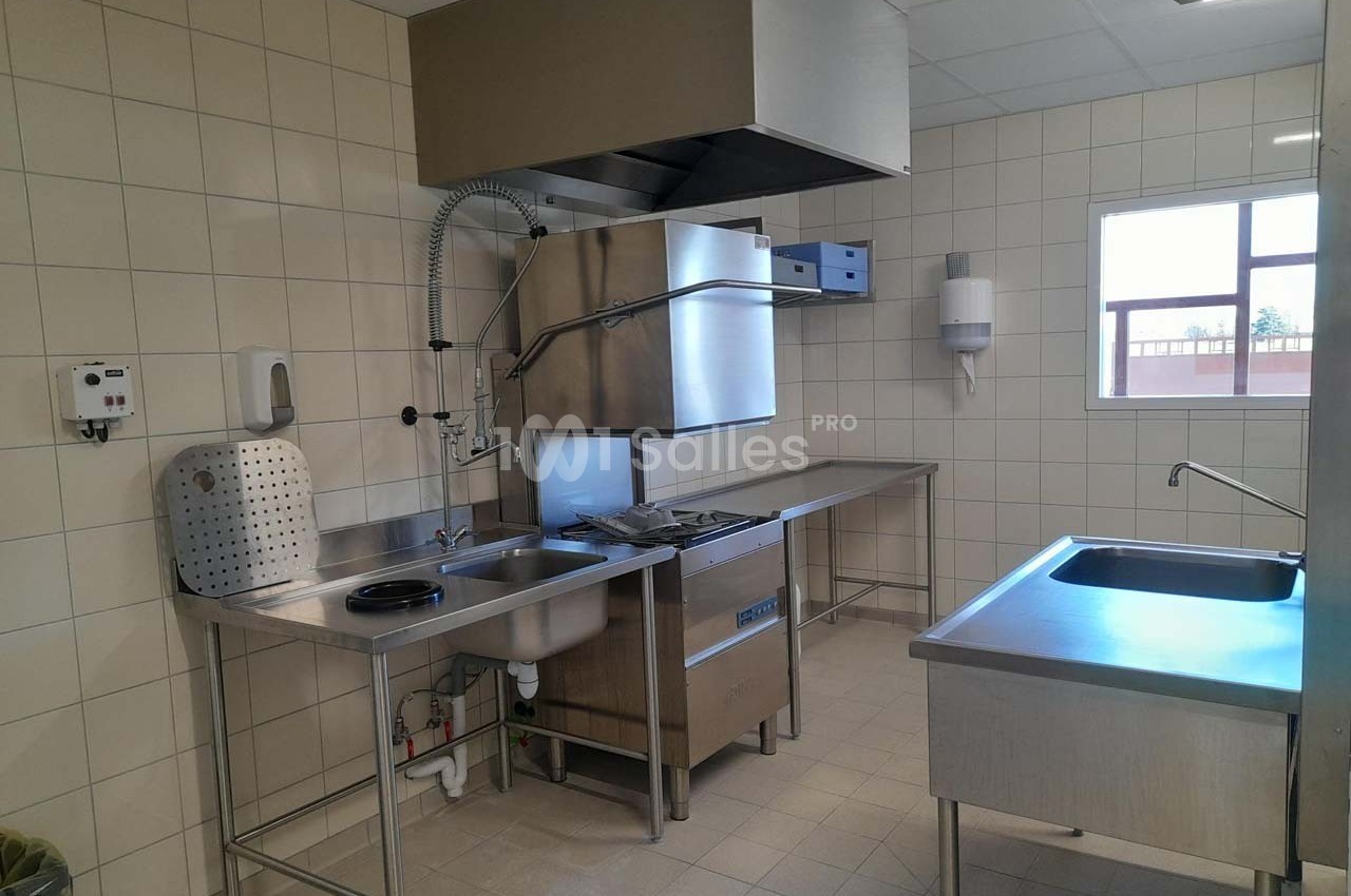 Cuisine professionnelle avec évier, lave-vaisselle industriel, plans de travail en inox et carrelage mural beige.