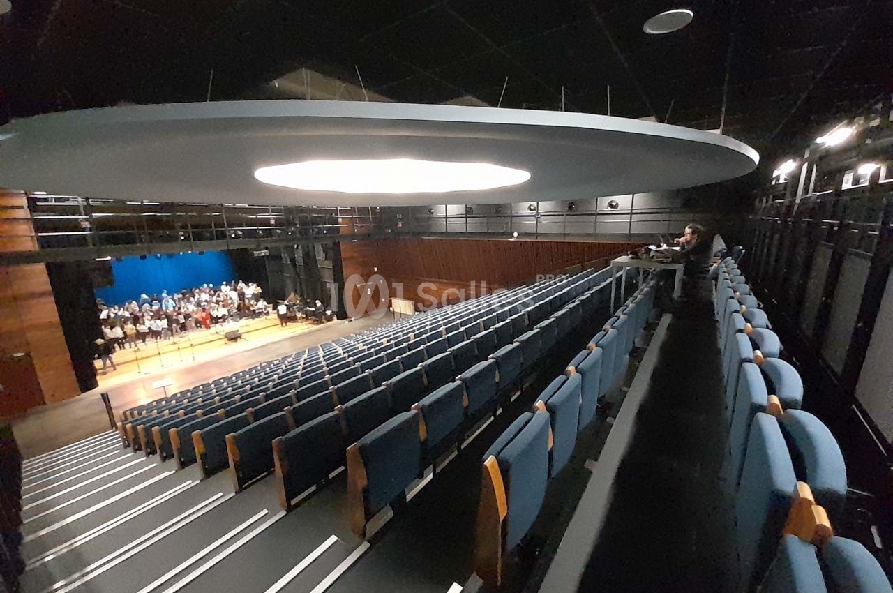 Salle de spectacle avec sièges bleus, scène éclairée en arrière-plan et quelques personnes présentes.