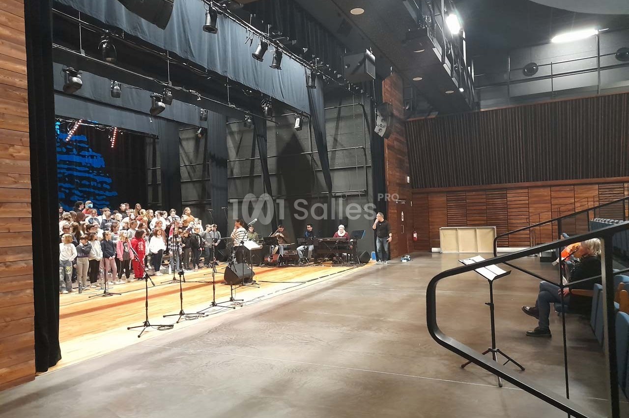 Un groupe de personnes répète sur une scène dans une salle de spectacle, avec un public restreint en arrière-plan.