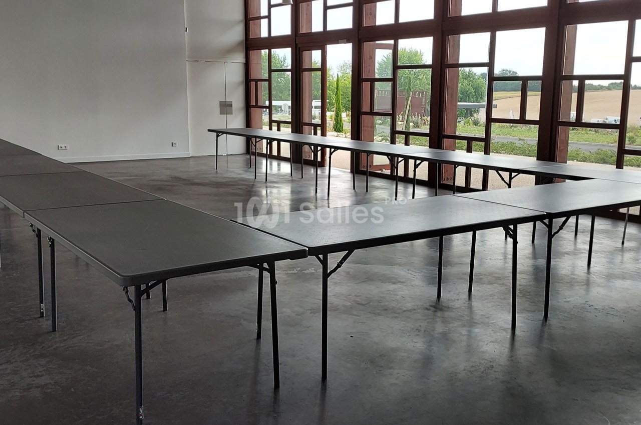 Salle lumineuse avec de grandes fenêtres, tables pliantes noires disposées en U sur un sol en béton.