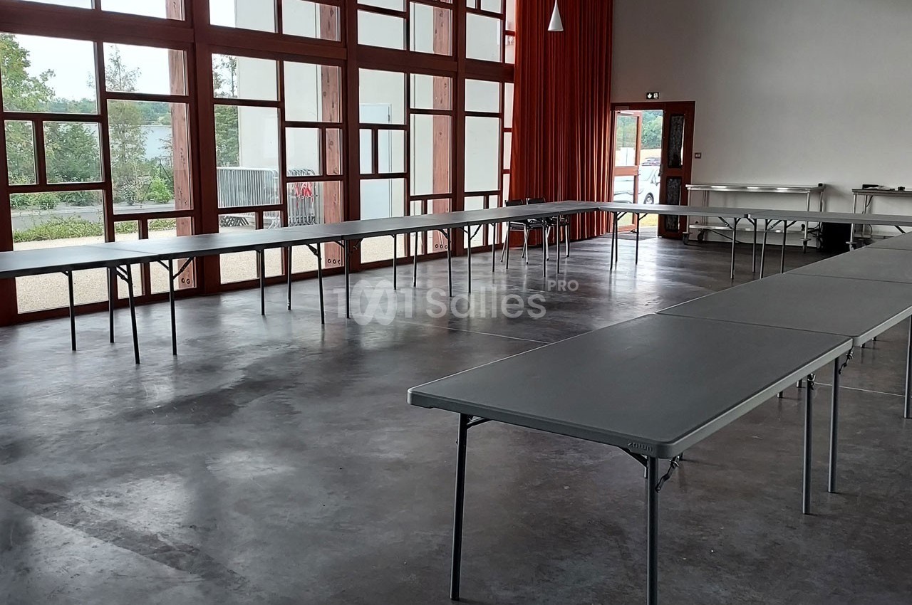 Salle lumineuse avec de grandes baies vitrées, tables pliantes grises disposées en U sur un sol en béton.