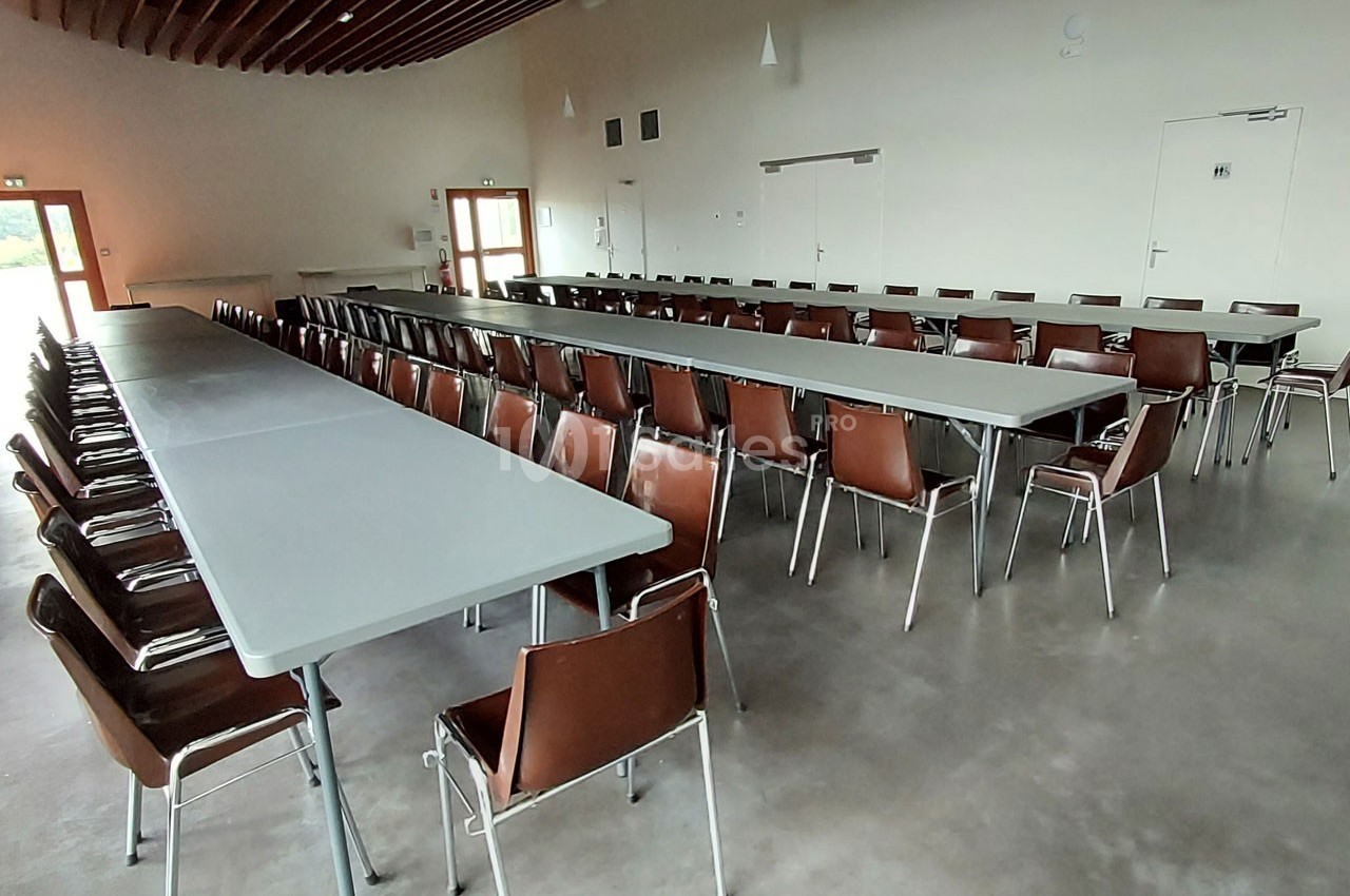 Salle avec de longues tables grises alignées et des chaises marron, éclairée par des fenêtres et des luminaires suspendus.