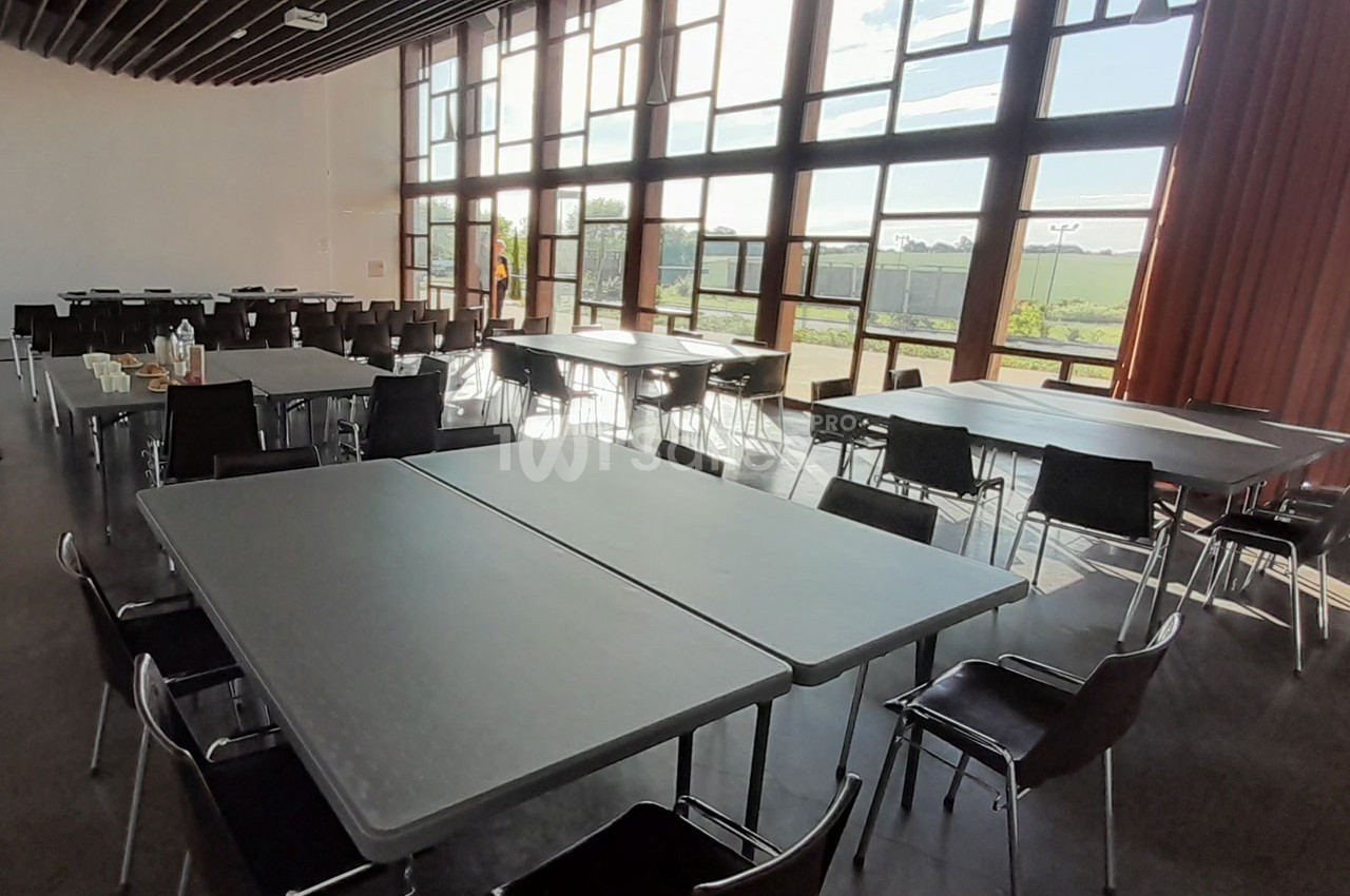 Salle lumineuse avec de grandes baies vitrées, tables rectangulaires grises et chaises noires disposées pour un événement.