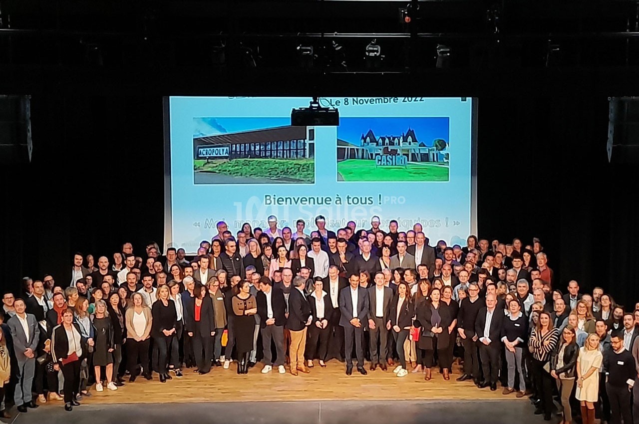 Photo de groupe sur une scène, avec un écran en arrière-plan affichant ’Bienvenue à tous !’ et des images de bâtiments.