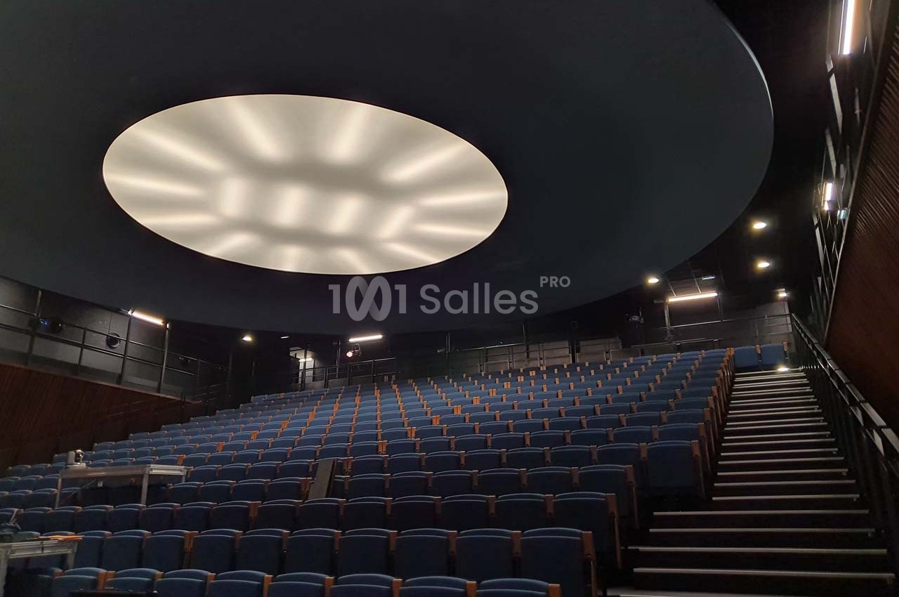 Salle de conférence vide avec des rangées de sièges bleus et un plafond circulaire éclairé au centre.