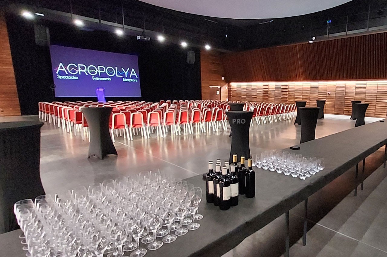 Salle de conférence avec rangées de chaises rouges, scène éclairée et table préparée avec verres et bouteilles de vin.