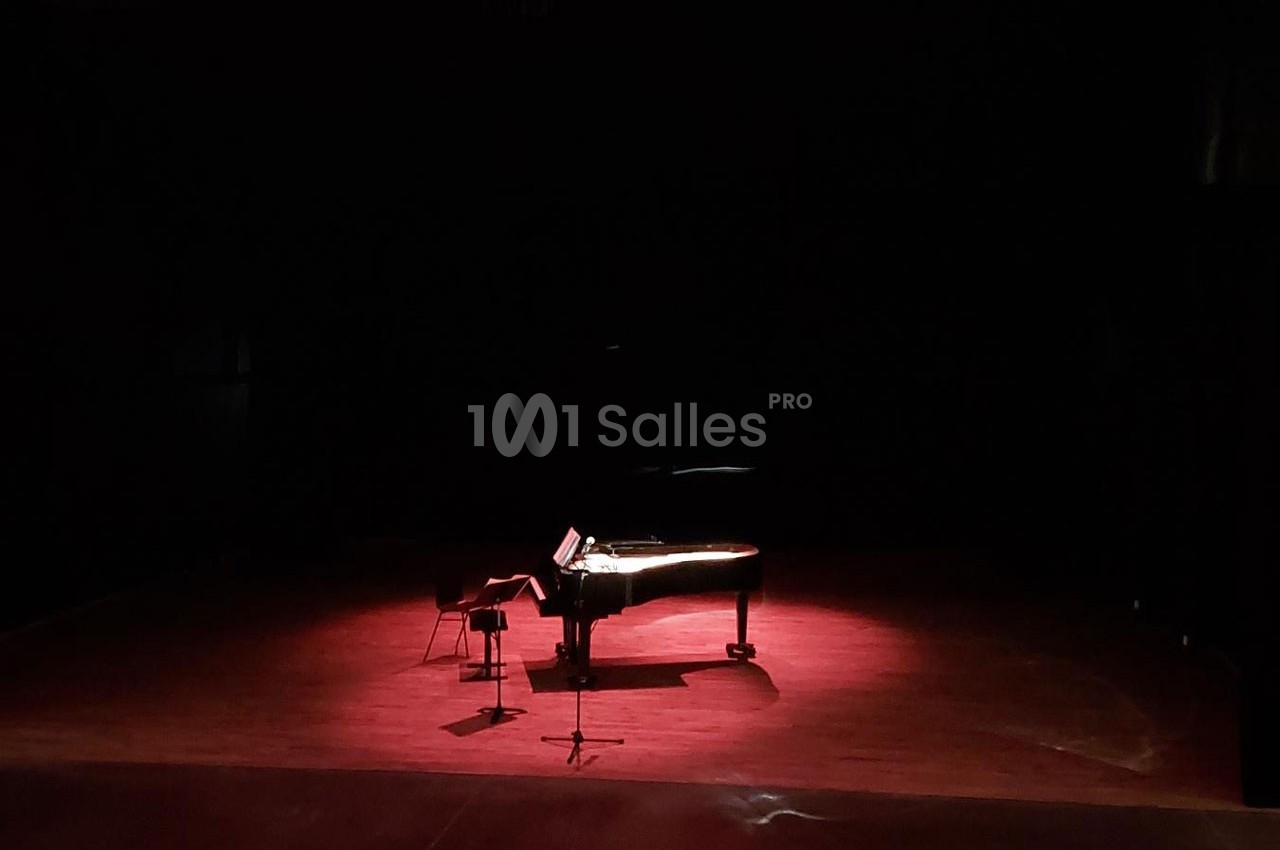 Un piano à queue éclairé par une lumière rouge sur une scène sombre, avec un tabouret et un pupitre.
