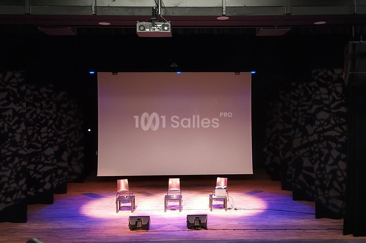 Trois chaises alignées sur une scène en bois avec un écran de projection blanc en arrière-plan.