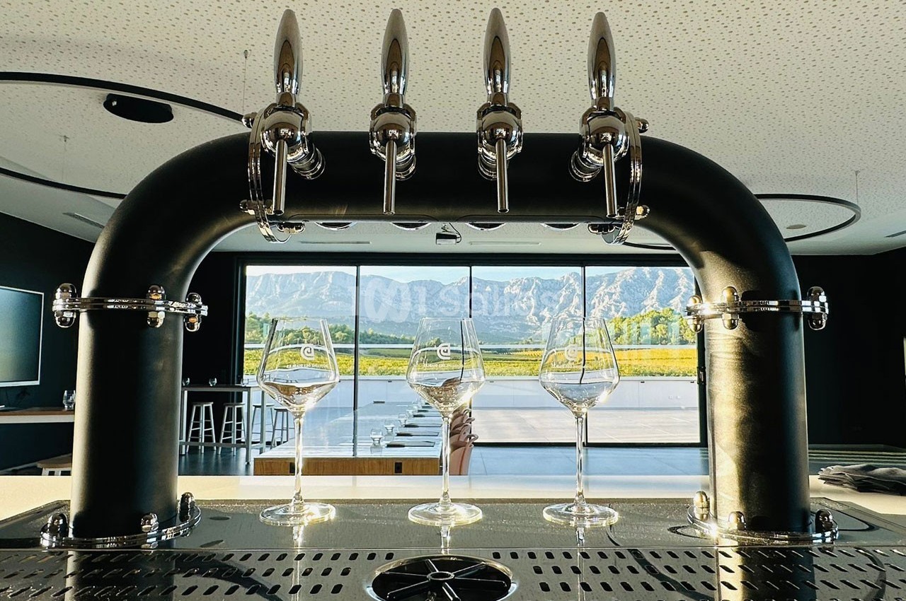 Quatre verres à vin alignés sous des robinets de bière, avec vue sur des montagnes et des vignes à travers une baie vitrée.
