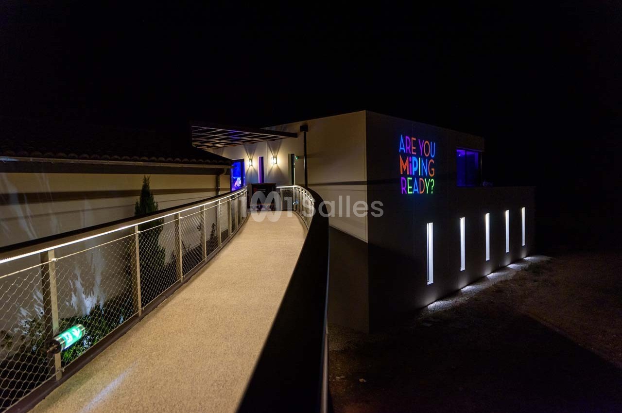Passerelle éclairée menant à un bâtiment moderne avec des lumières colorées et un message lumineux sur la façade.