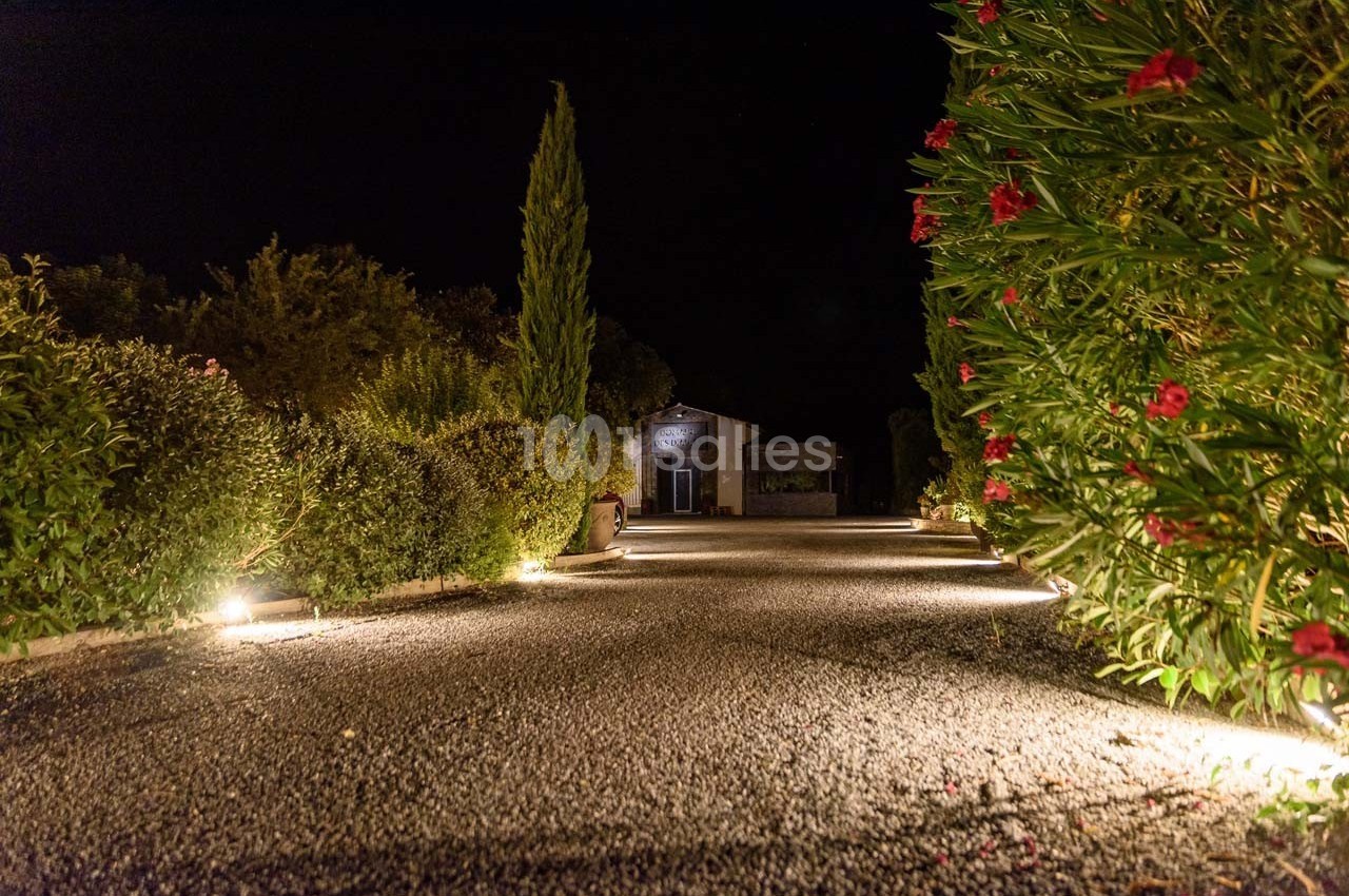 Allée de gravier éclairée par des lumières au sol, bordée de buissons et menant à une maison en pierre, de nuit.