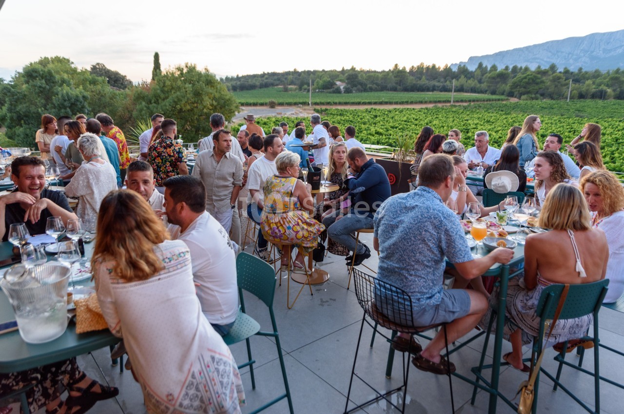 Groupe de personnes dînant en terrasse avec vue sur des vignobles et des collines au coucher du soleil.
