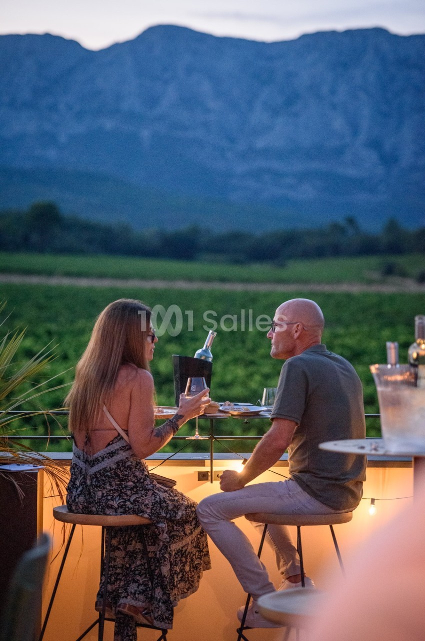 Un couple discute autour d'un verre de vin en terrasse au coucher du soleil, avec des montagnes en arrière-plan.