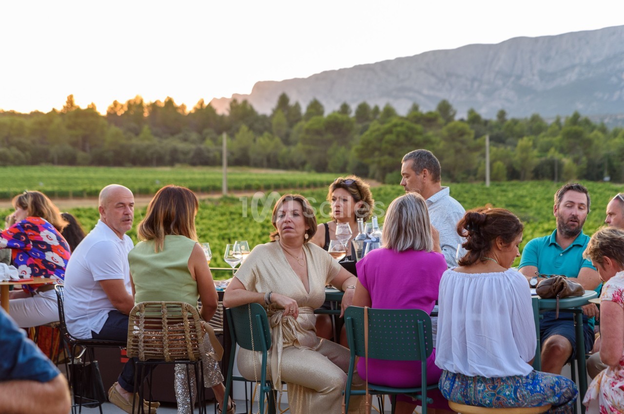 Des personnes assises à une table en extérieur, discutant et partageant un repas devant un paysage de vignobles au coucher…