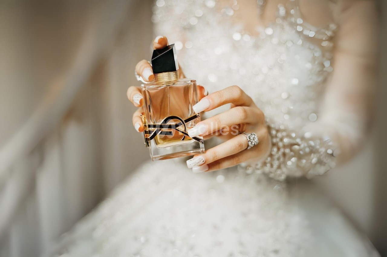 Main tenant un flacon de parfum élégant avec un design doré, en arrière-plan une robe ornée de perles.
