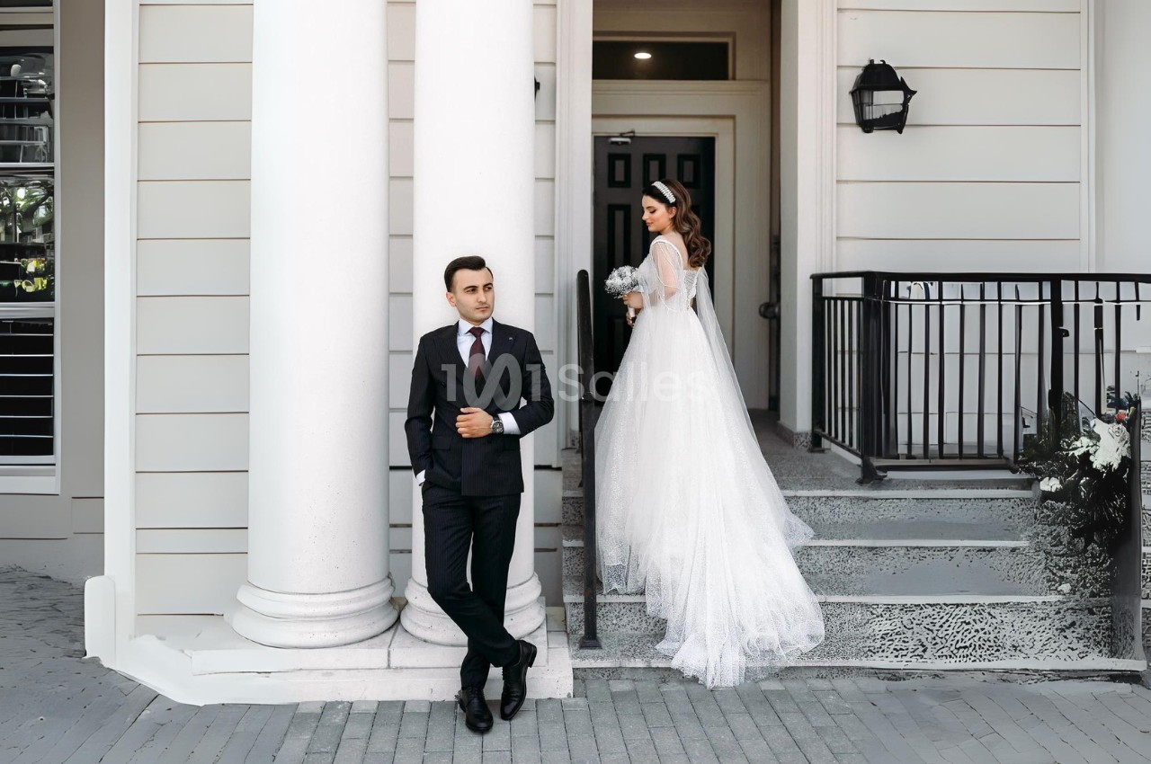 Un homme en costume se tient devant un bâtiment, une femme en robe de mariée descend les marches derrière lui.