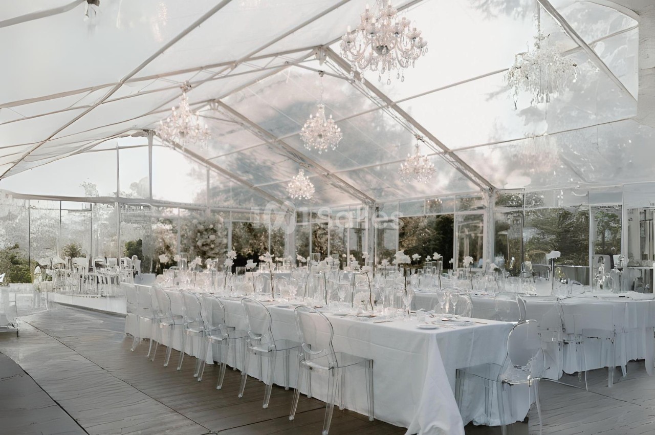 Salle de réception sous une tente transparente avec tables dressées, chaises transparentes et lustres suspendus.