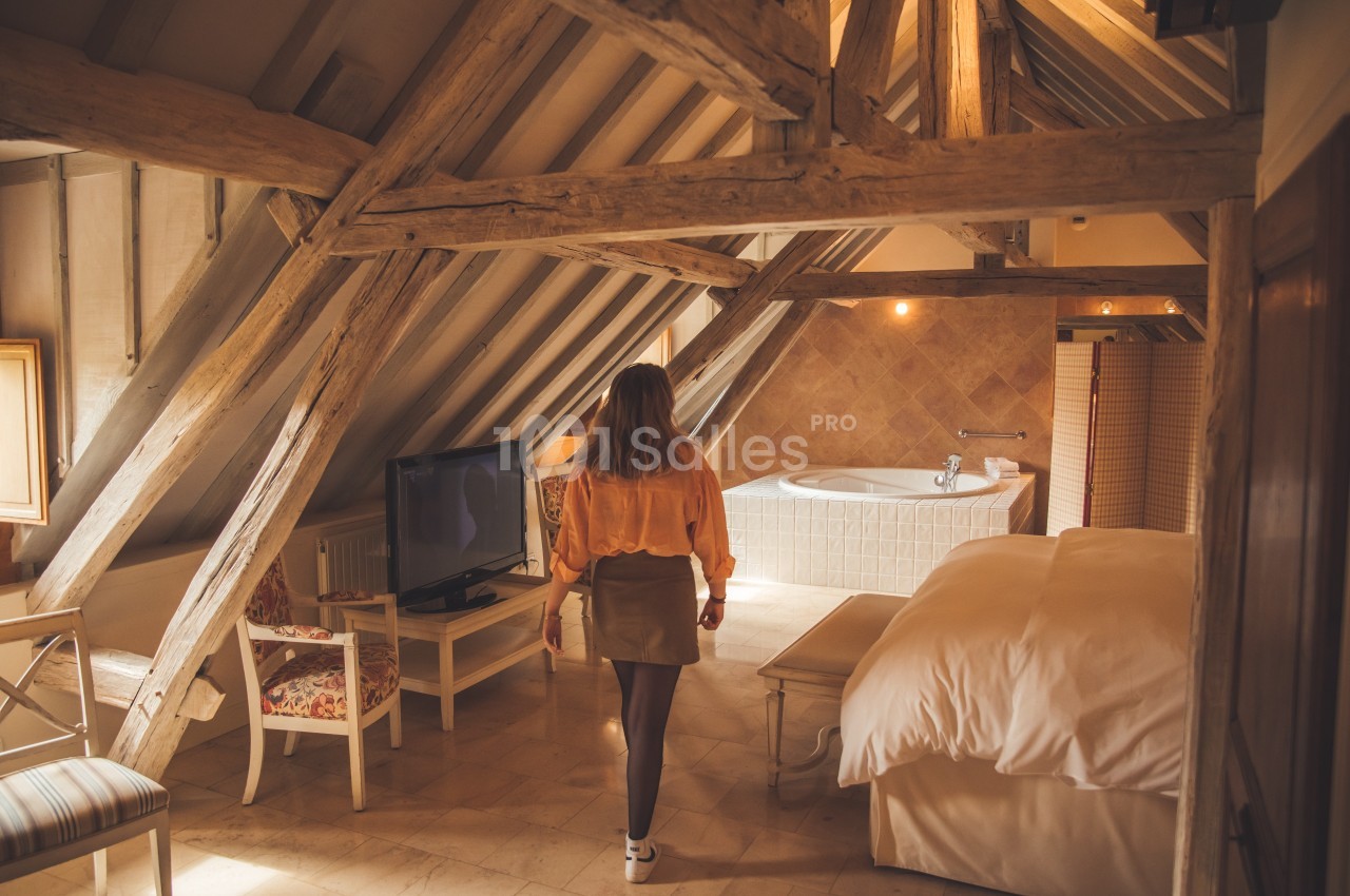 Femme de dos dans une chambre mansardée avec poutres apparentes, lit, télévision et baignoire en arrière-plan.