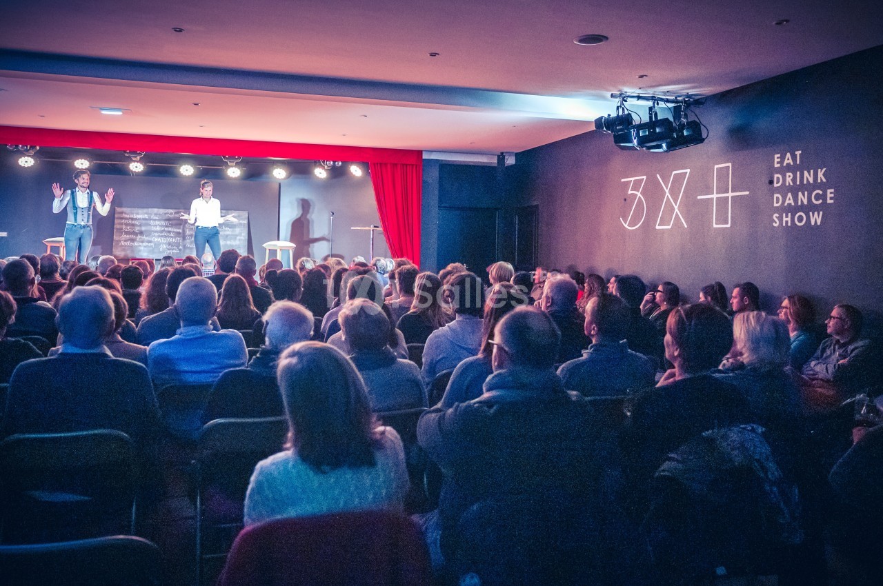 Salle de spectacle avec un public assis face à deux personnes sur scène, éclairée par des projecteurs.