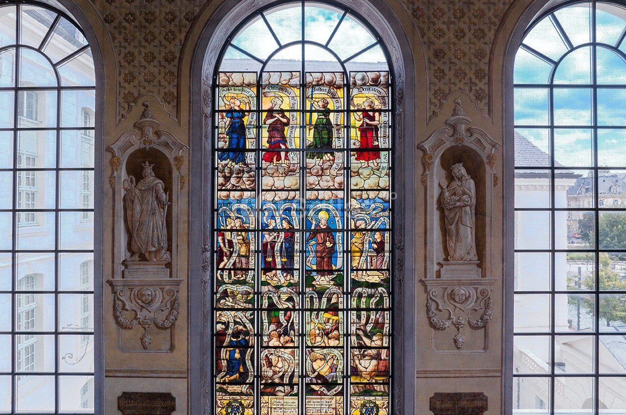 Vitrail coloré représentant des scènes religieuses, encadré par deux statues et des fenêtres laissant entrer la lumière.