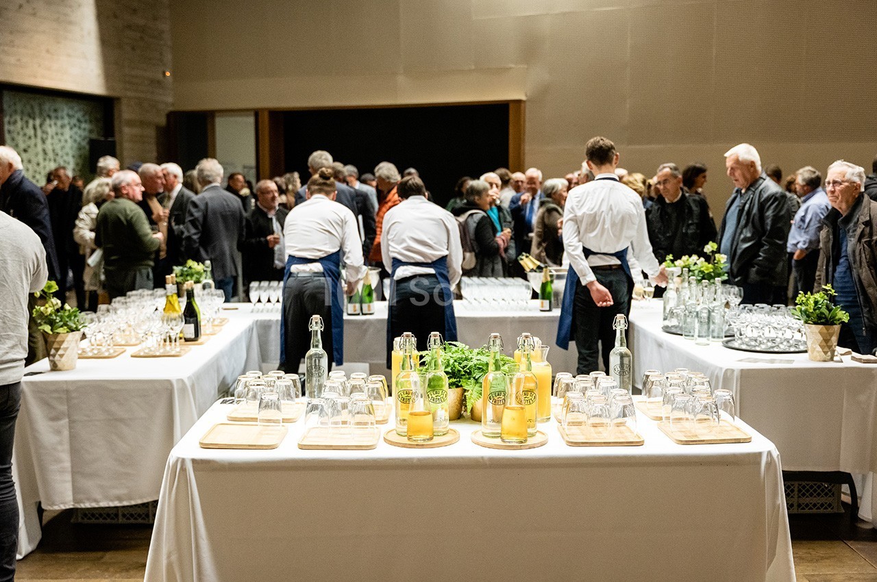 Des tables dressées avec boissons et verres lors d'un événement rassemblant des invités dans une salle.