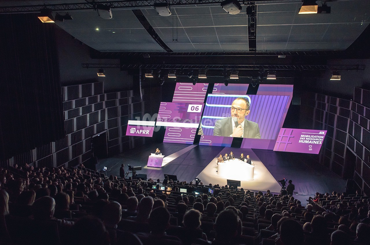 Conférence dans un auditorium avec un intervenant sur scène, écran géant et public assis dans une salle sombre.