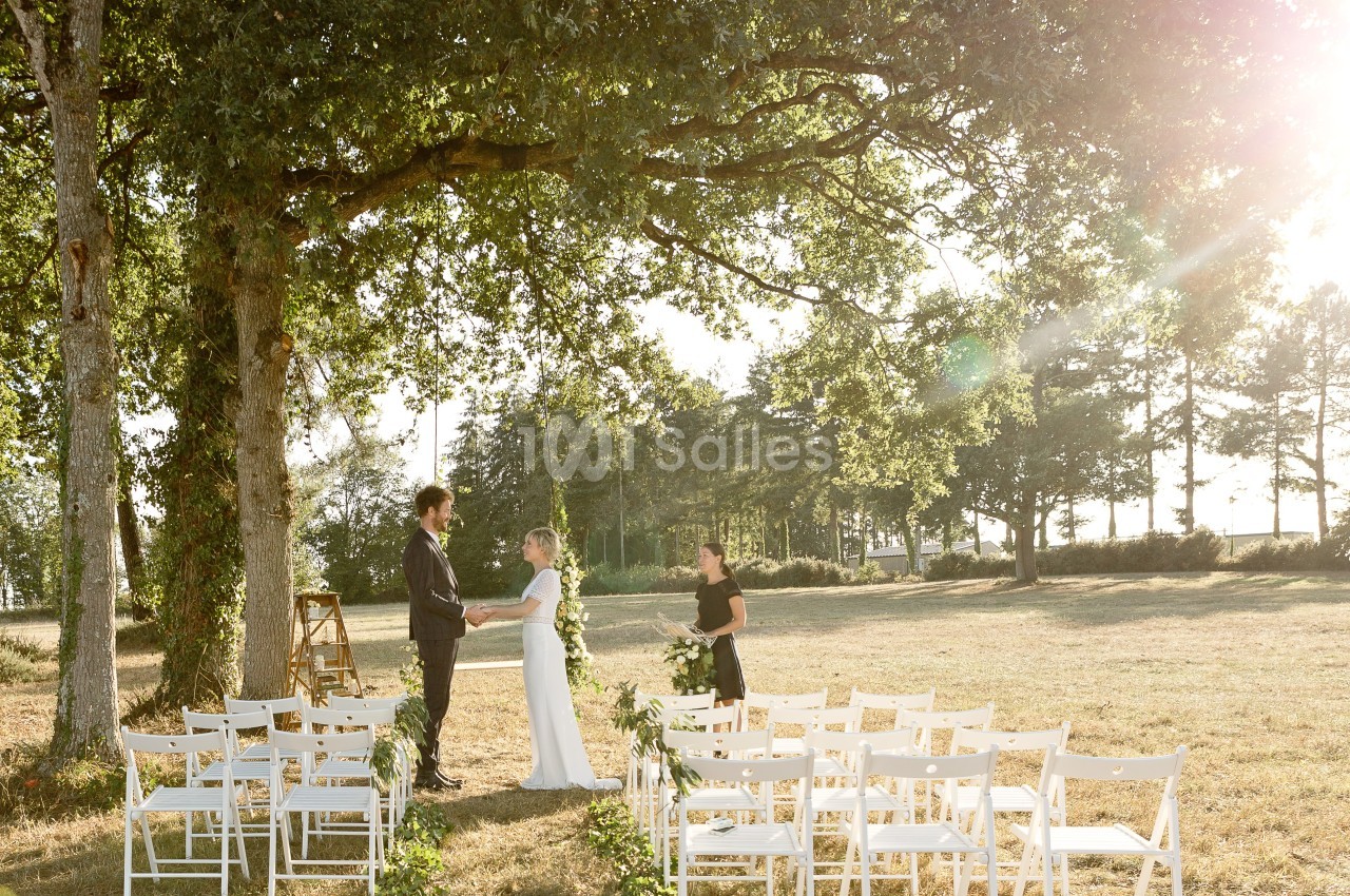 Un couple échange ses vœux sous un grand arbre dans un cadre champêtre, avec des chaises blanches disposées en allée.