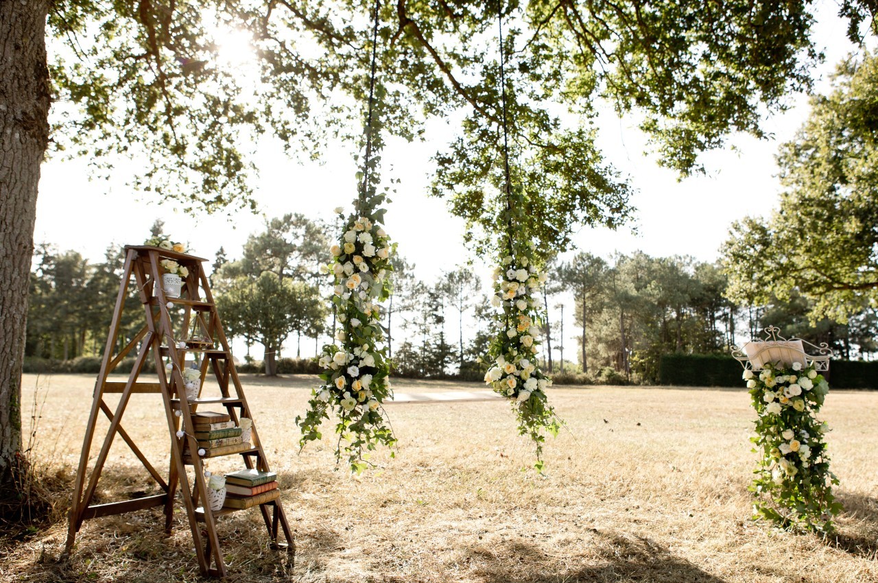 Trois balançoires décorées de fleurs blanches et feuillages suspendues à un arbre dans un champ ensoleillé.