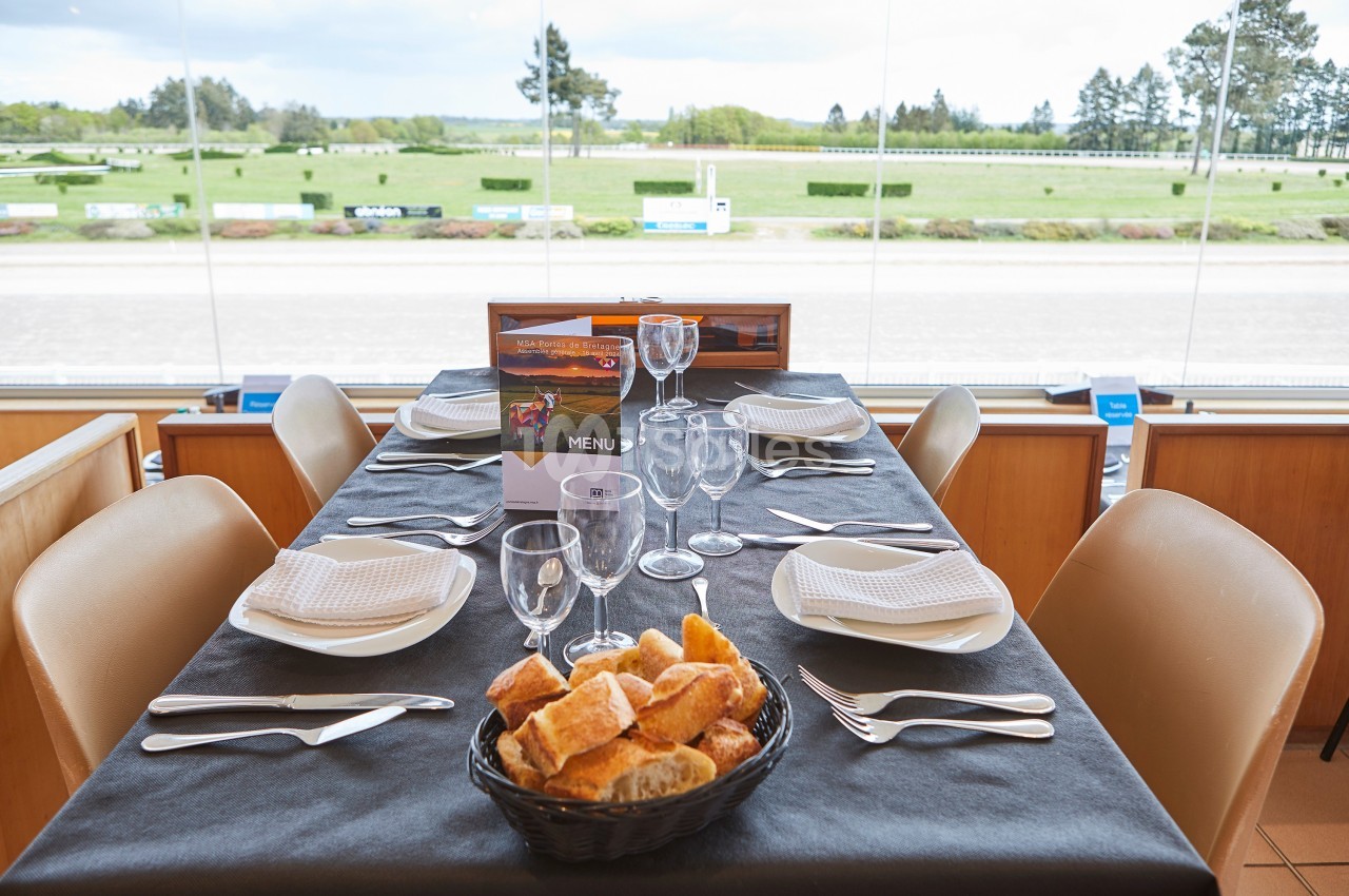 Table dressée avec vaisselle et panier de pain, située dans une salle avec vue sur un hippodrome et des espaces verts.