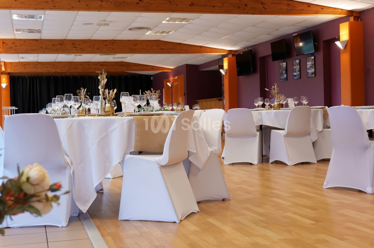 Salle de réception décorée avec des tables dressées, nappes blanches et chaises recouvertes, éclairage chaleureux.