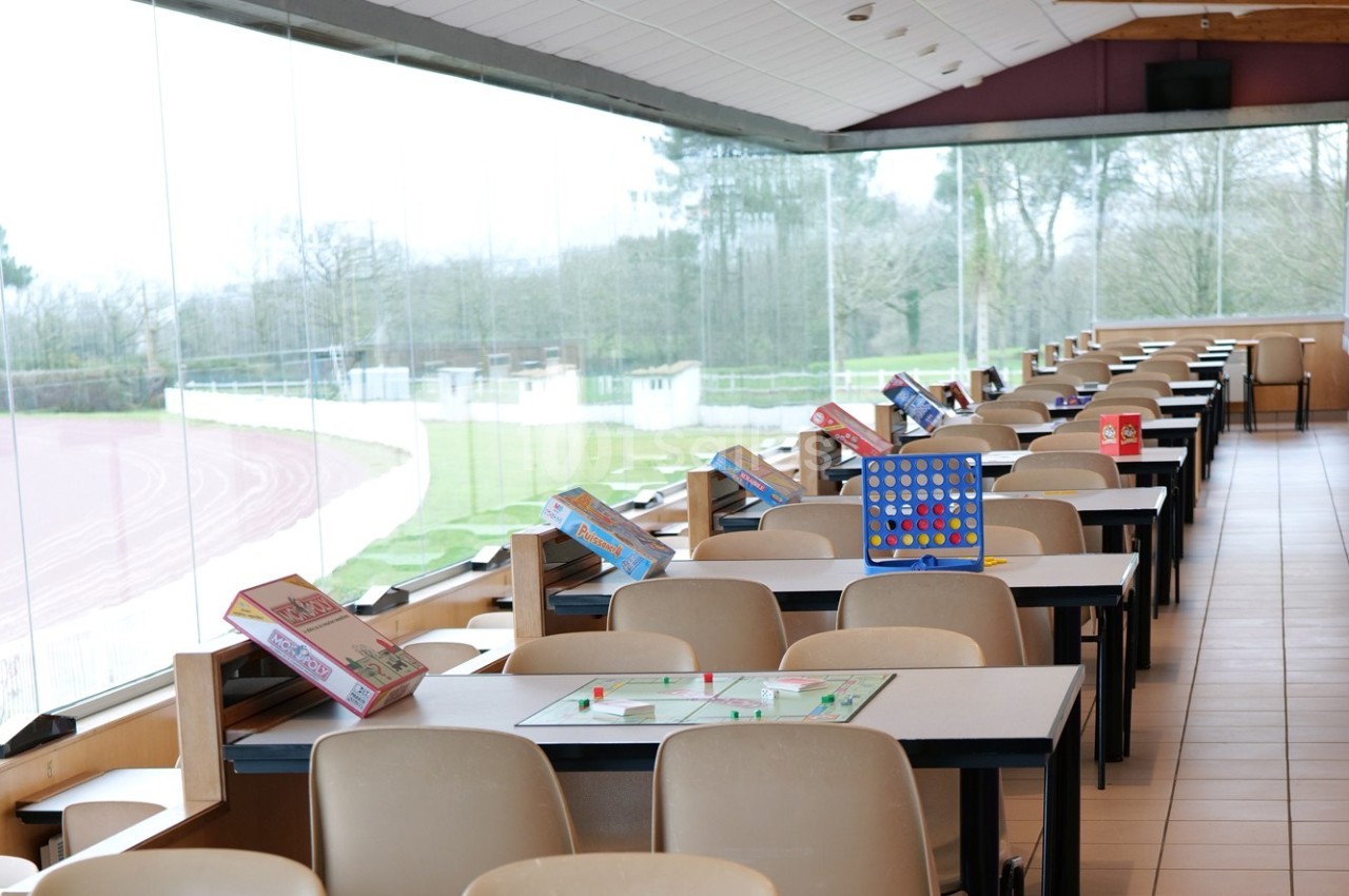 Salle lumineuse avec des tables alignées, chacune équipée de jeux de société, vue sur une piste extérieure.