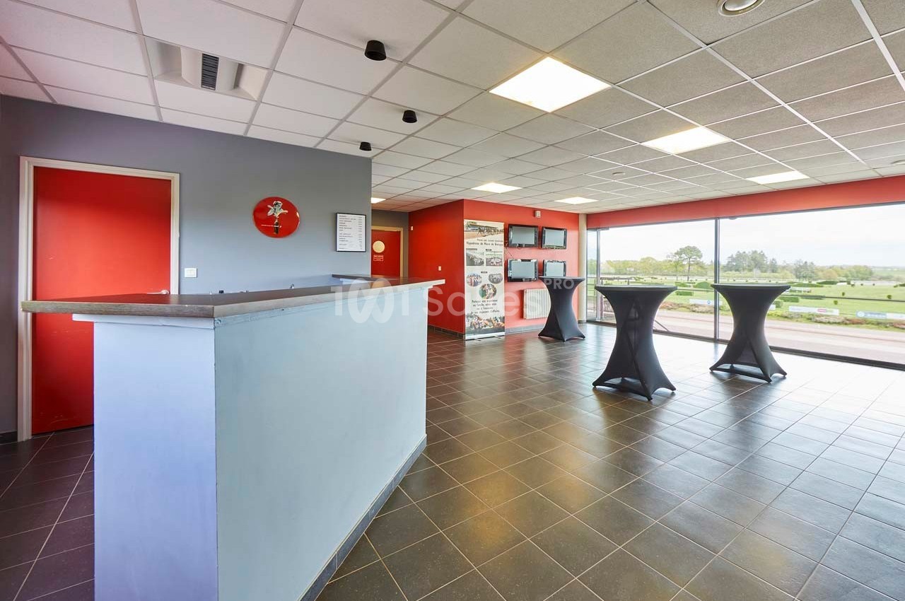 Salle lumineuse avec comptoir d'accueil, tables hautes noires et grande baie vitrée donnant sur un espace extérieur.