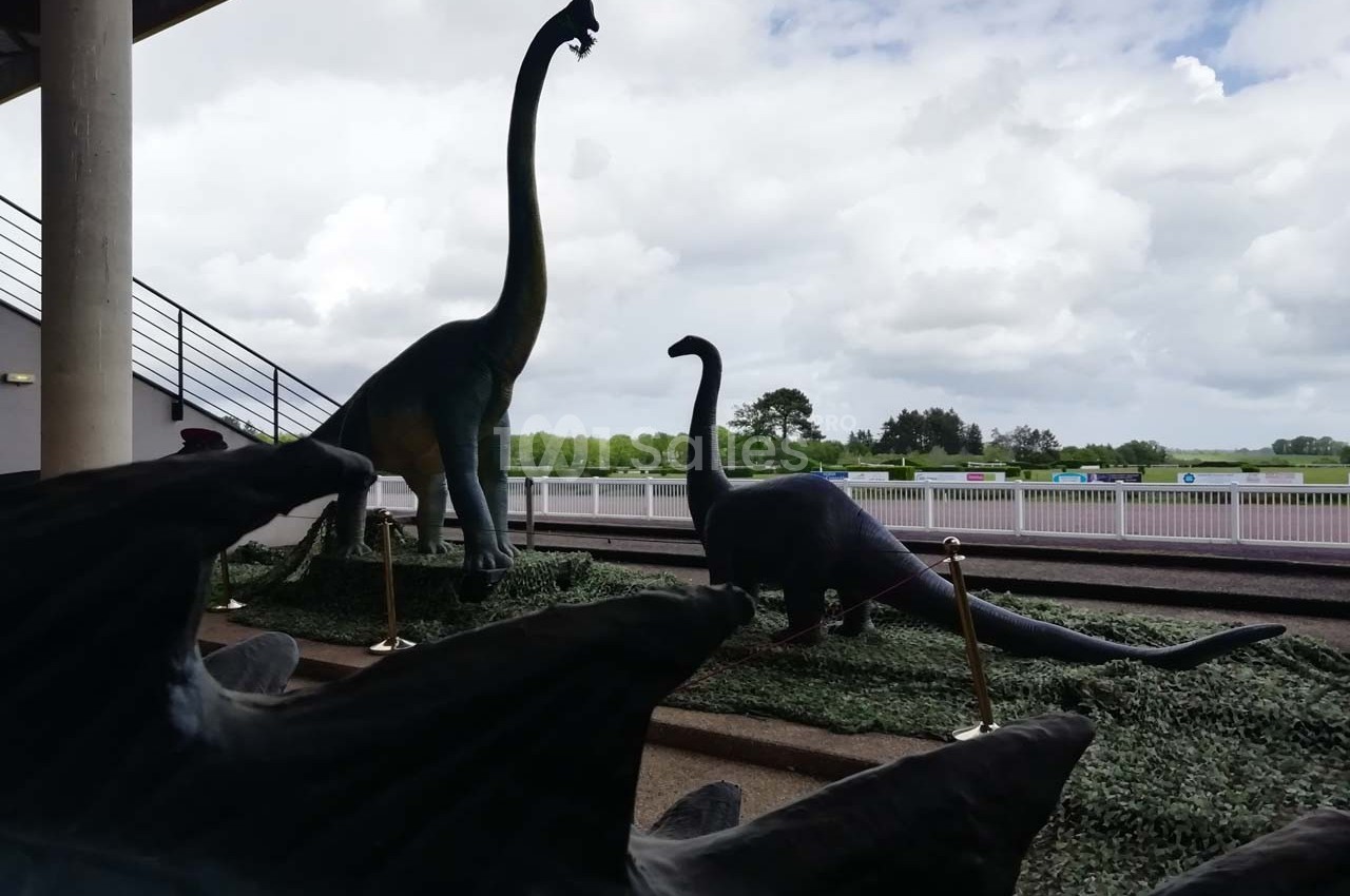 Statues de dinosaures herbivores sur un espace extérieur, avec un ciel nuageux et une barrière en arrière-plan.