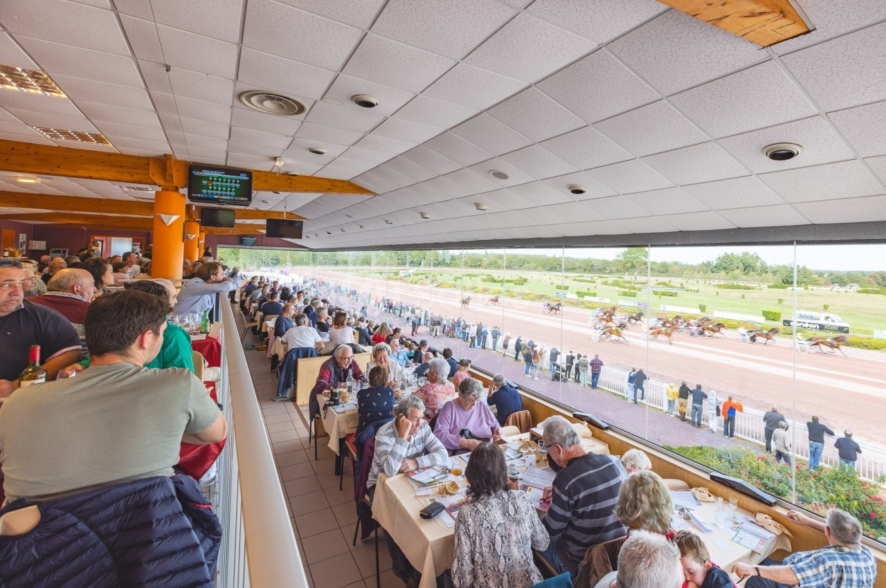 Vue intérieure d'un restaurant surplombant une piste de course hippique, avec des spectateurs et des chevaux en compétition.