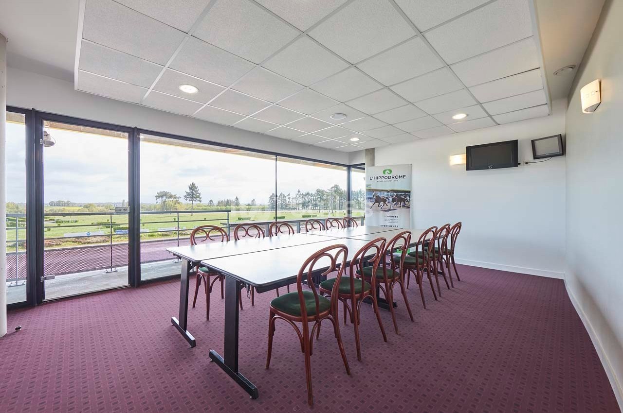 Salle de réunion lumineuse avec table rectangulaire, chaises en bois, grandes baies vitrées et vue sur un espace extérieur.