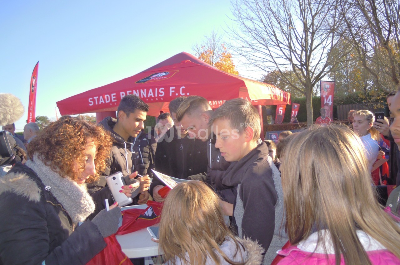 Des personnes rassemblées autour d'une table pour des autographes devant un stand du Stade Rennais F.C. en extérieur.