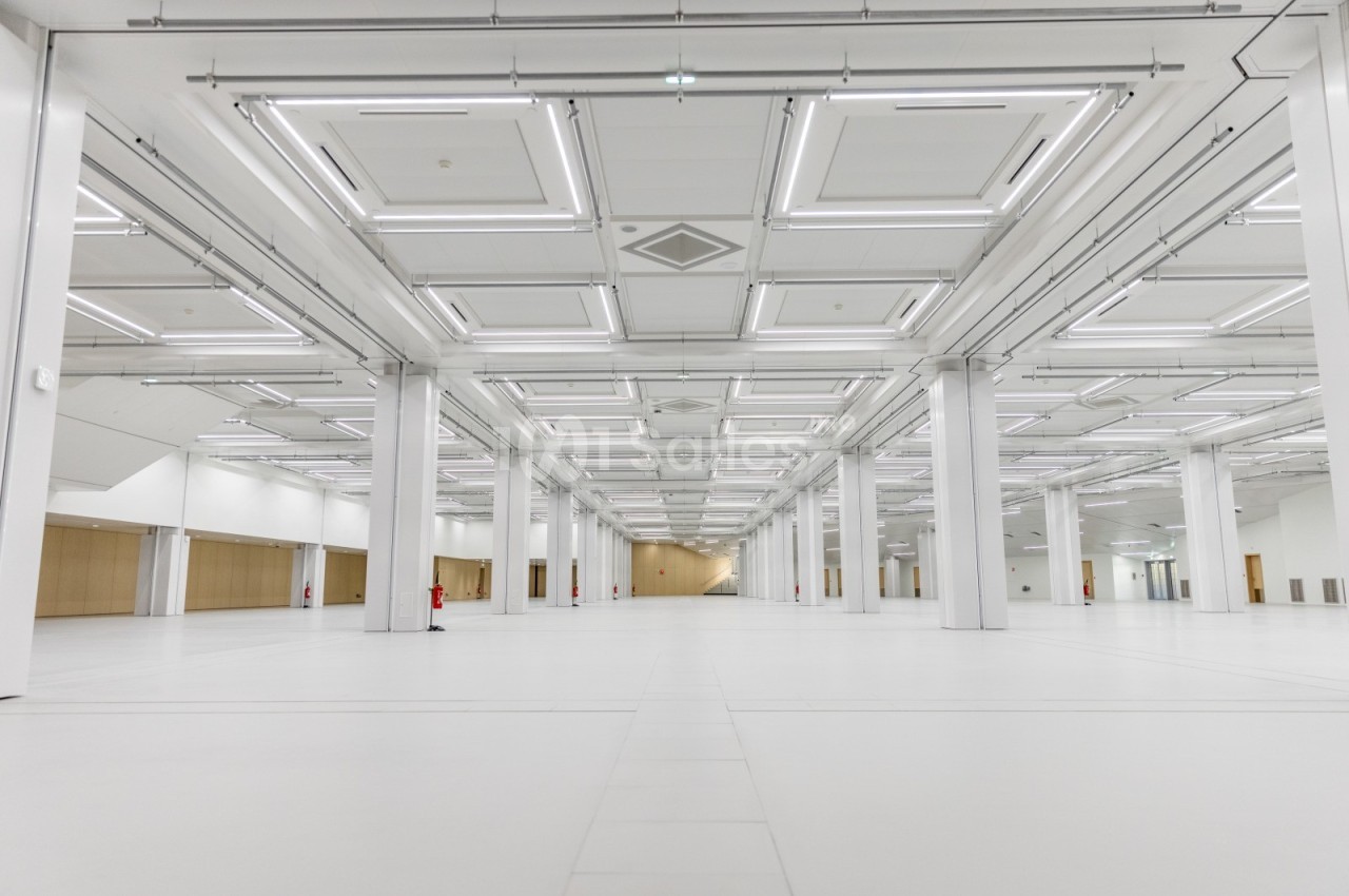 Grand espace intérieur vide avec un sol blanc, des colonnes et un plafond équipé de nombreux luminaires rectangulaires.