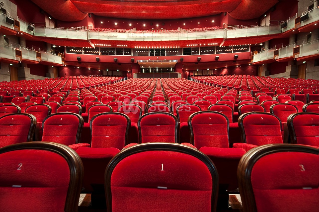 Salle de théâtre vide avec des rangées de sièges rouges alignés face à une scène éclairée.