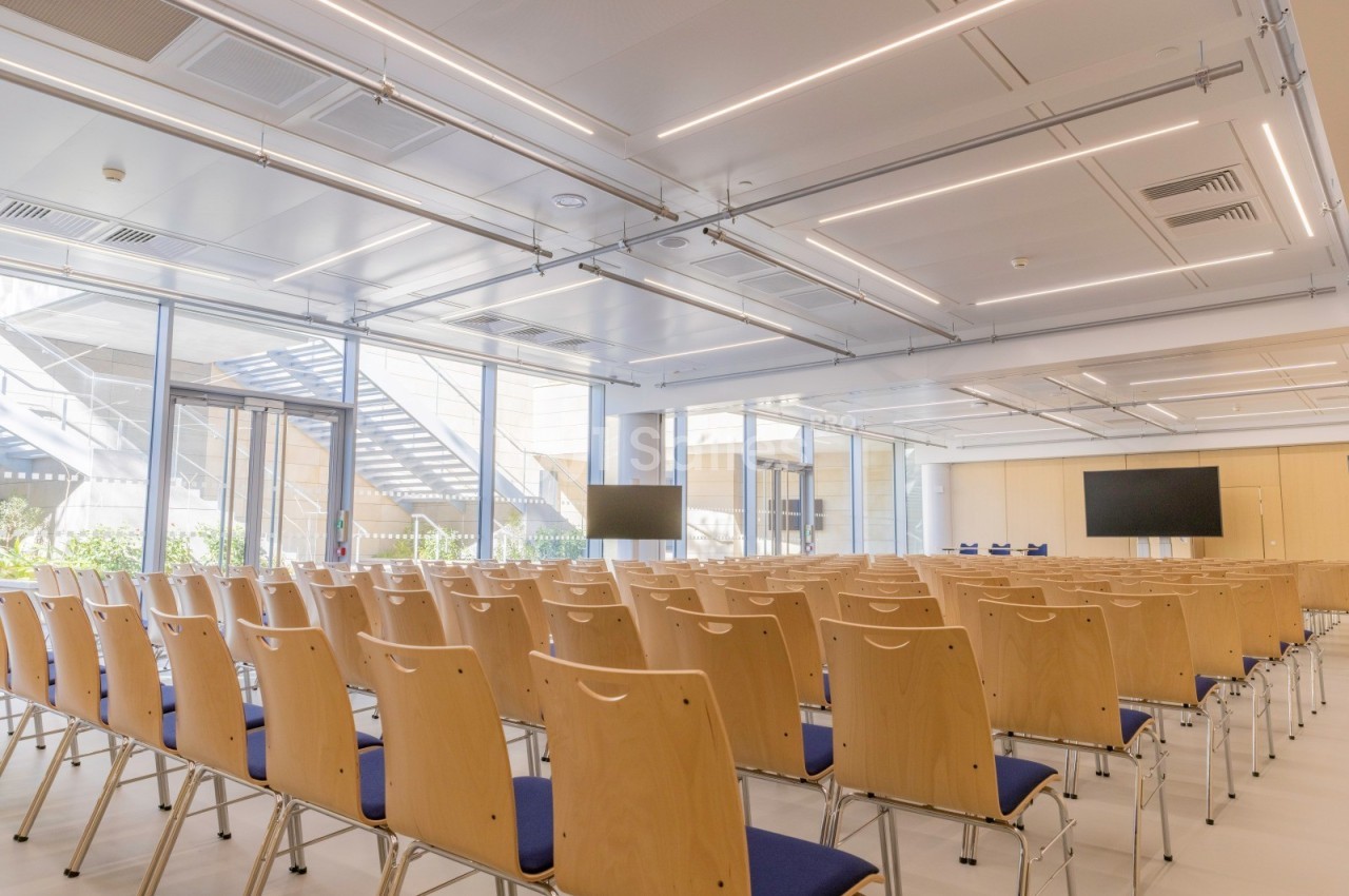 Salle de conférence lumineuse avec des rangées de chaises en bois, deux écrans et de grandes fenêtres donnant sur l…