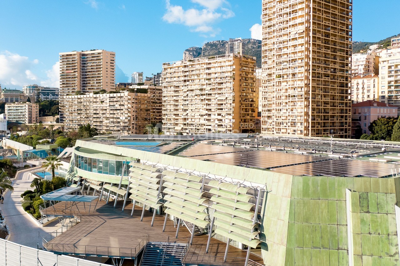 Vue d'un bâtiment moderne avec panneaux solaires sur le toit, entouré de hauts immeubles sous un ciel dégagé.