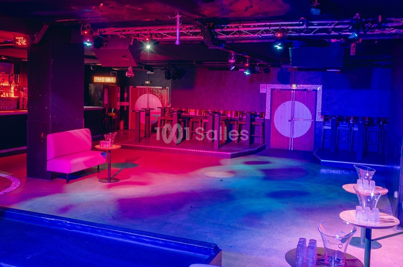 Salle de club vide avec éclairage coloré, banquette rose, tables basses et bar en arrière-plan.