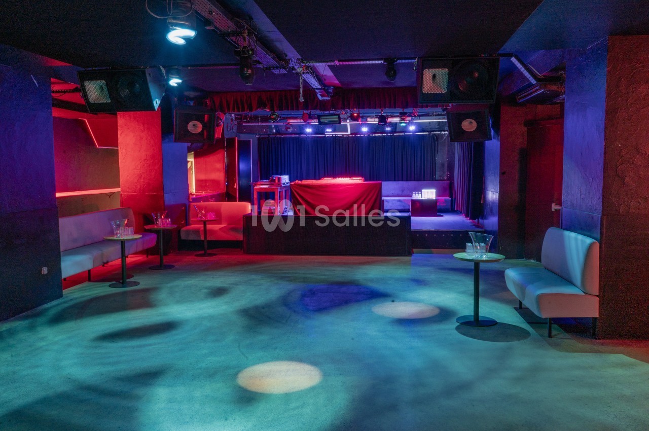 Salle de concert vide avec scène éclairée par des projecteurs colorés, entourée de banquettes et tables basses.