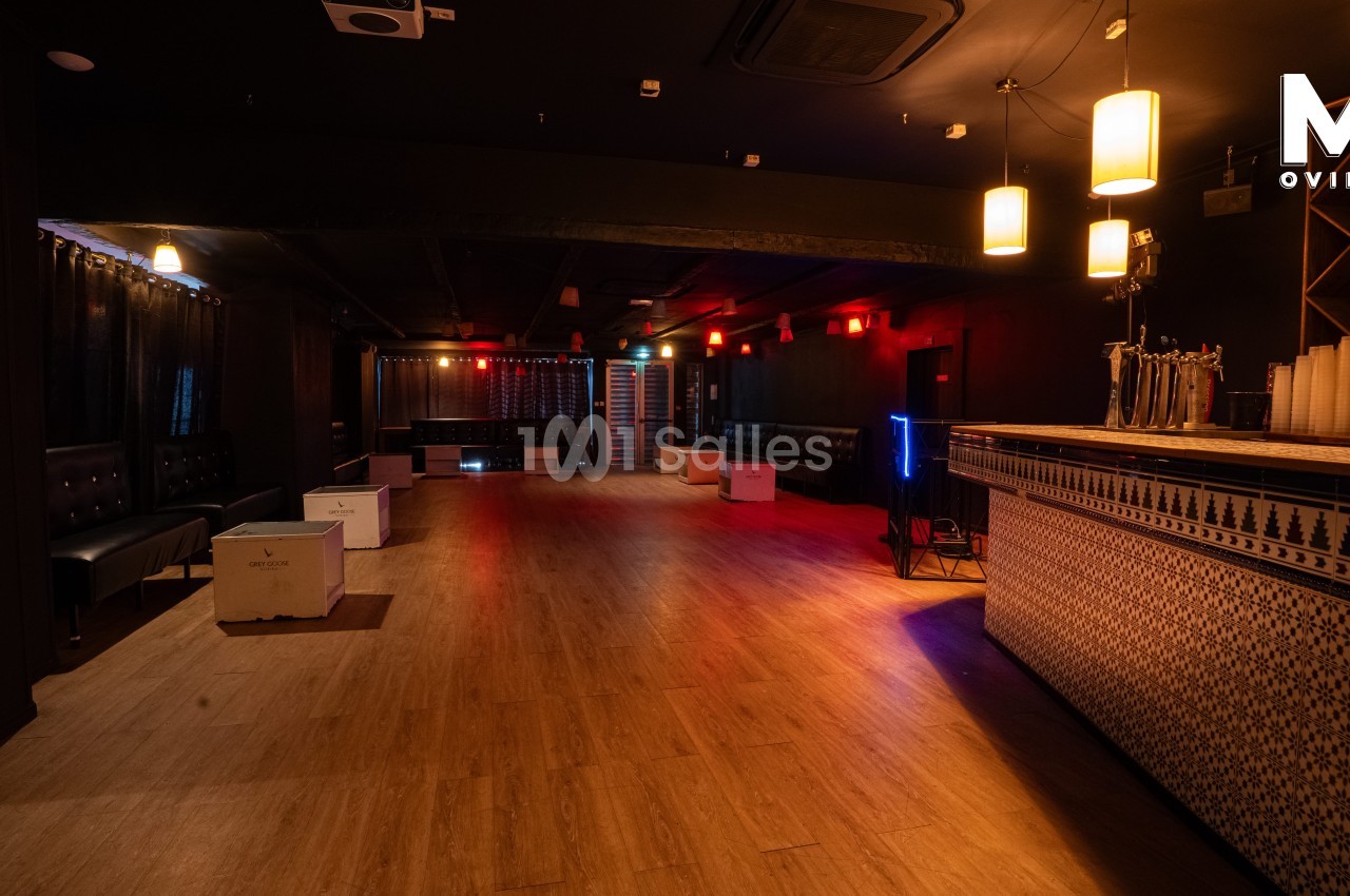 Salle sombre avec parquet, bar à droite, banquettes noires sur les côtés et éclairage tamisé.