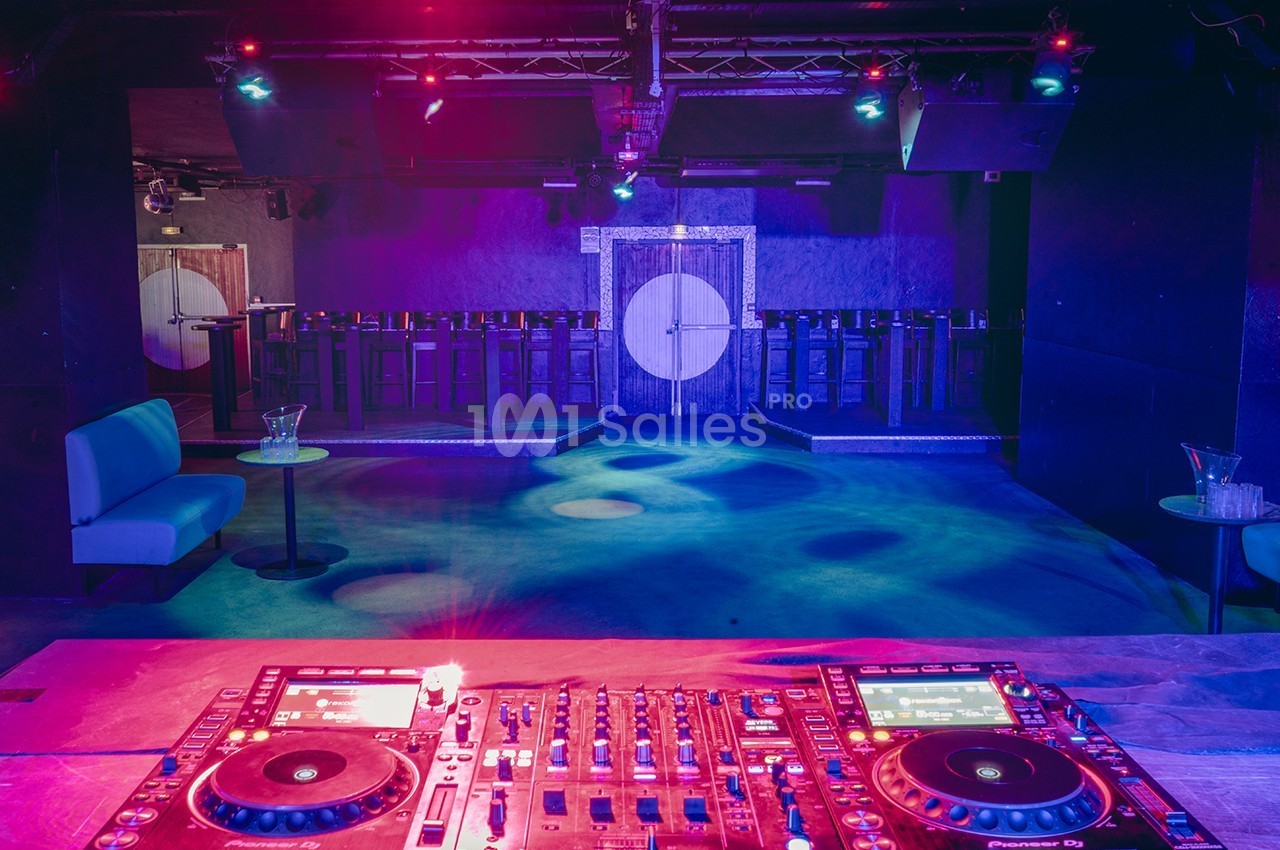 Salle de discothèque vide avec une console de DJ au premier plan, éclairée par des lumières colorées.