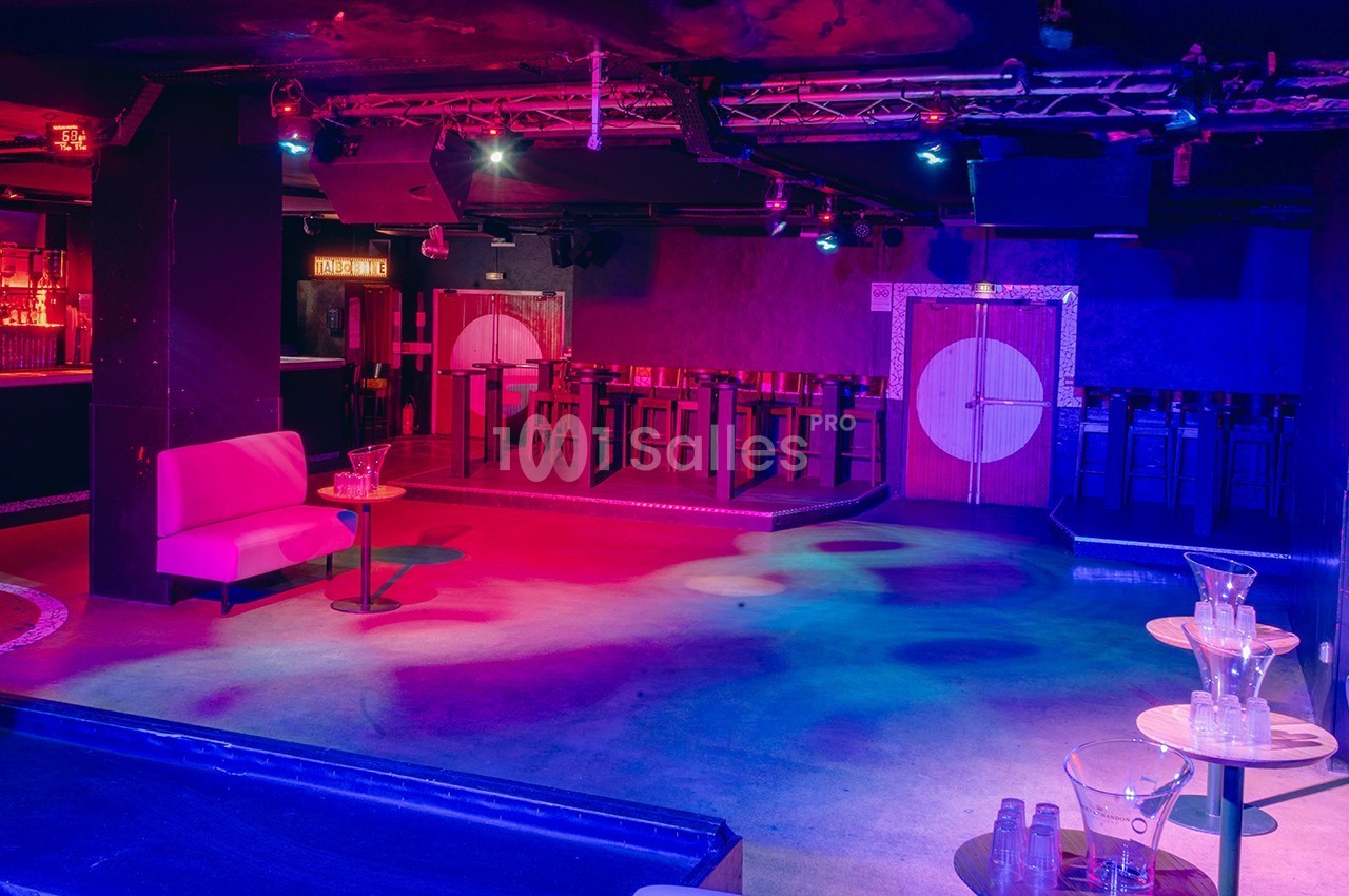 Salle de club vide avec éclairage coloré, banquette rose, tables basses et bar en arrière-plan.