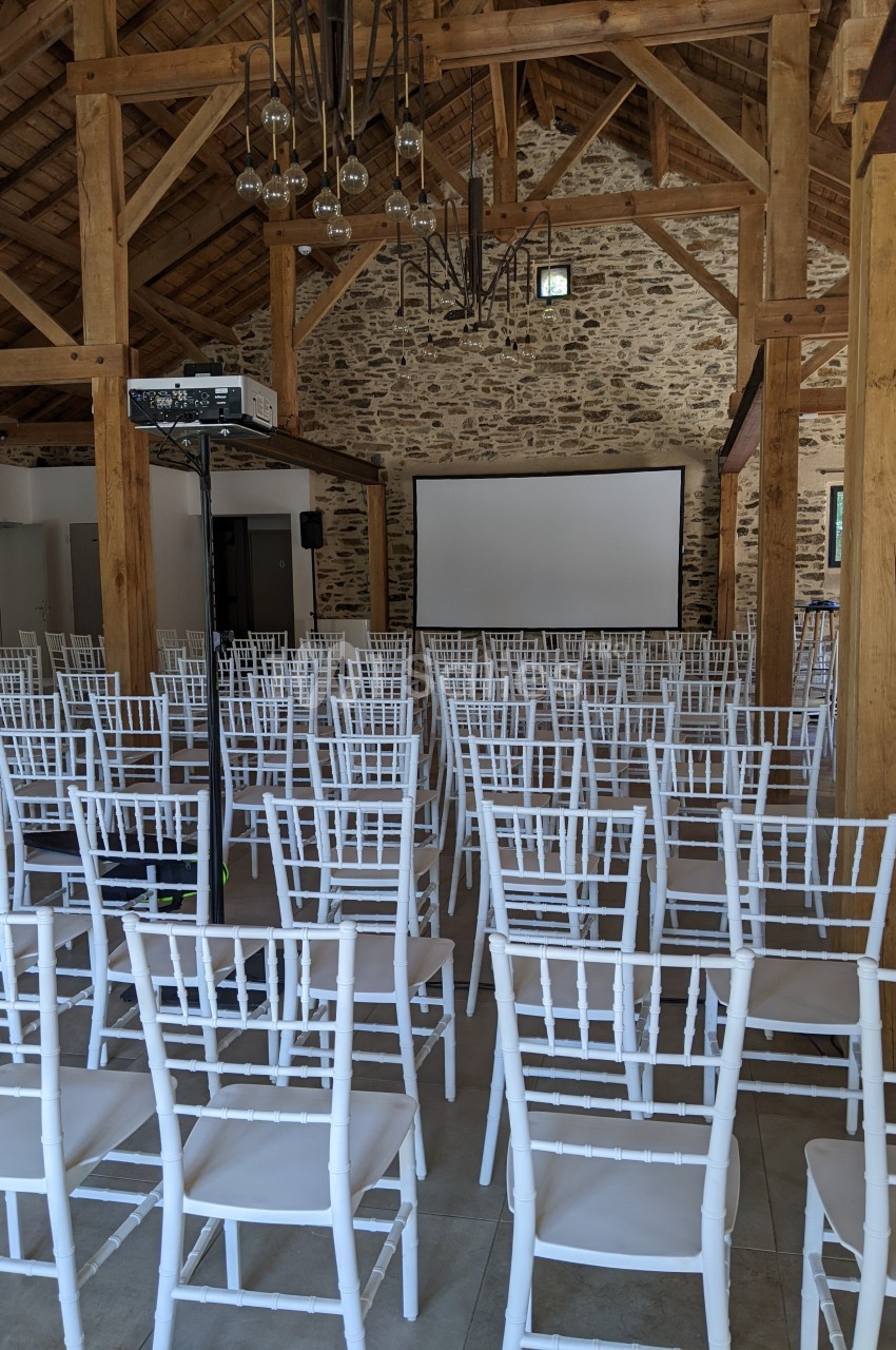 Salle de conférence avec des rangées de chaises blanches face à un écran de projection, sous une charpente en bois.