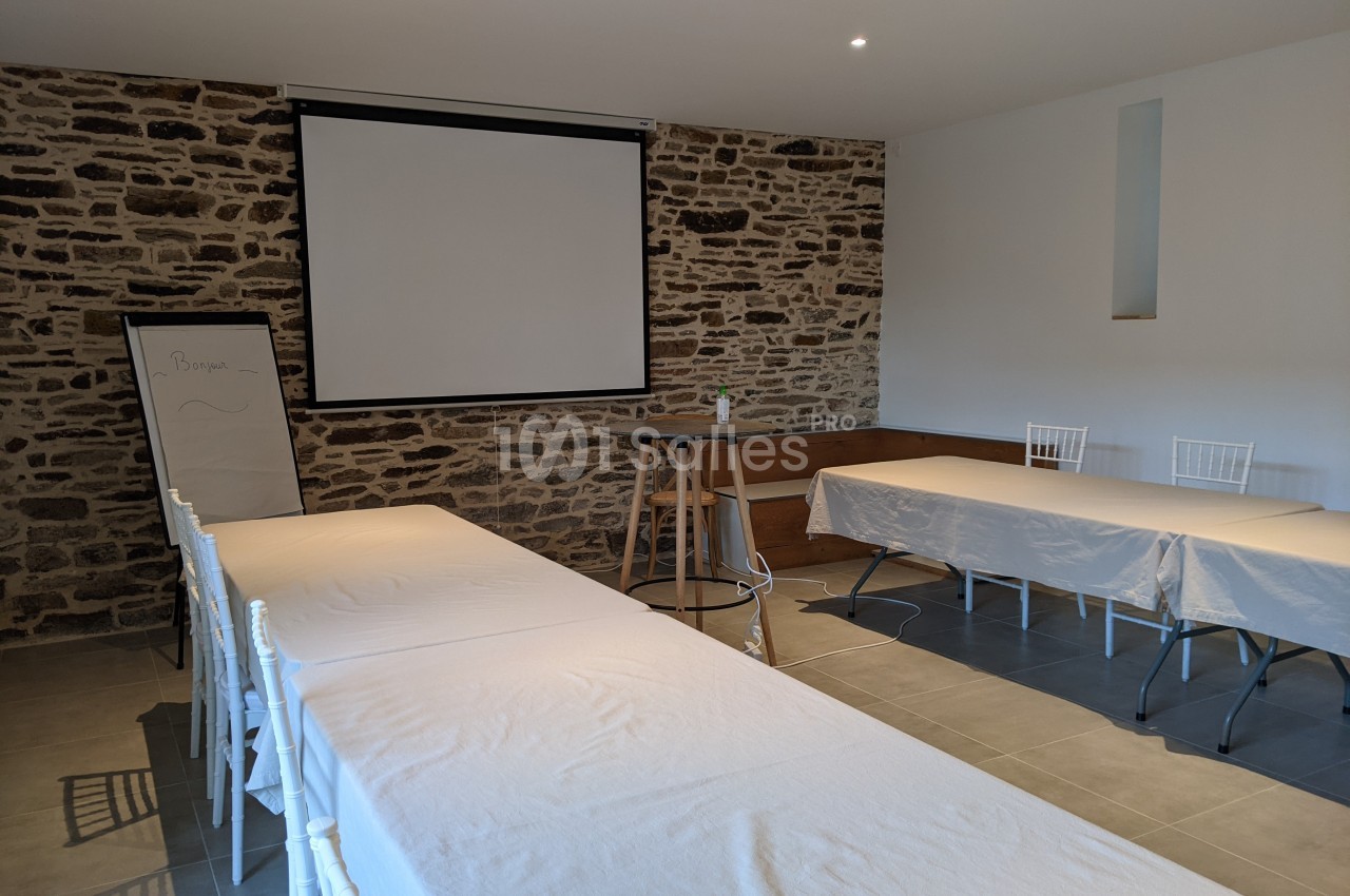 Salle de réunion avec murs en pierre, tables couvertes de nappes blanches, un tableau et un écran de projection.