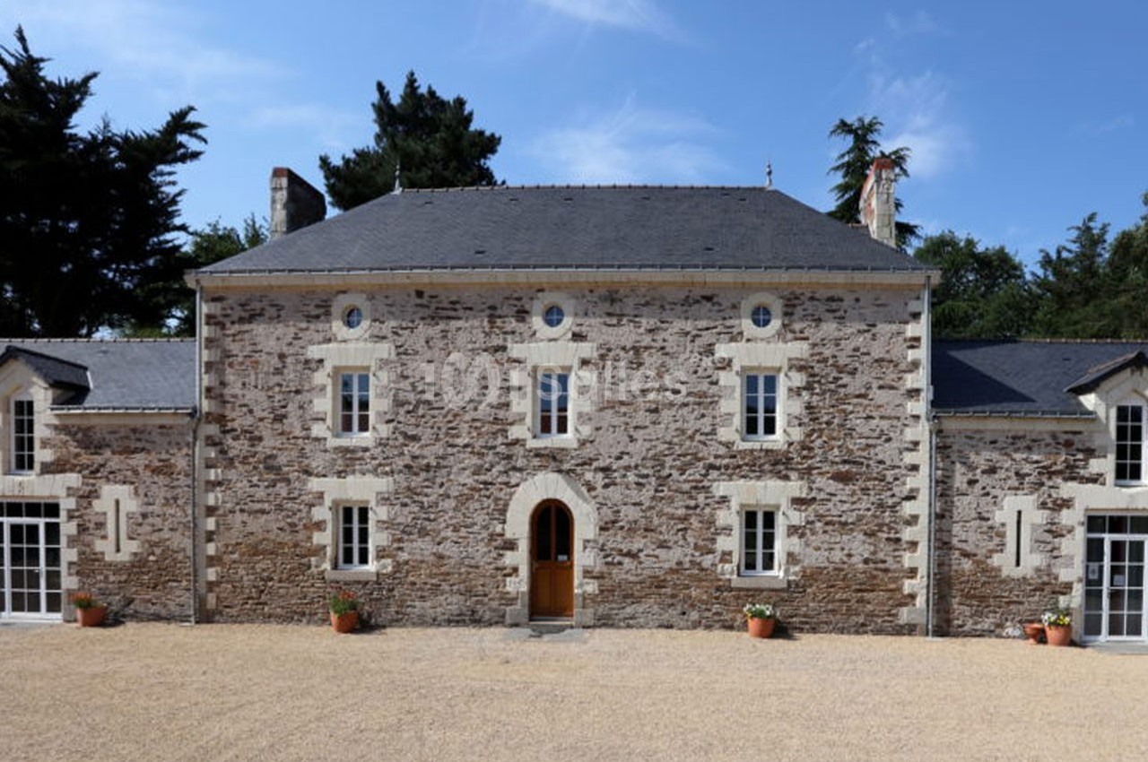 Façade d'une maison en pierre avec toit en ardoise, entourée d'arbres et donnant sur une cour en gravier.