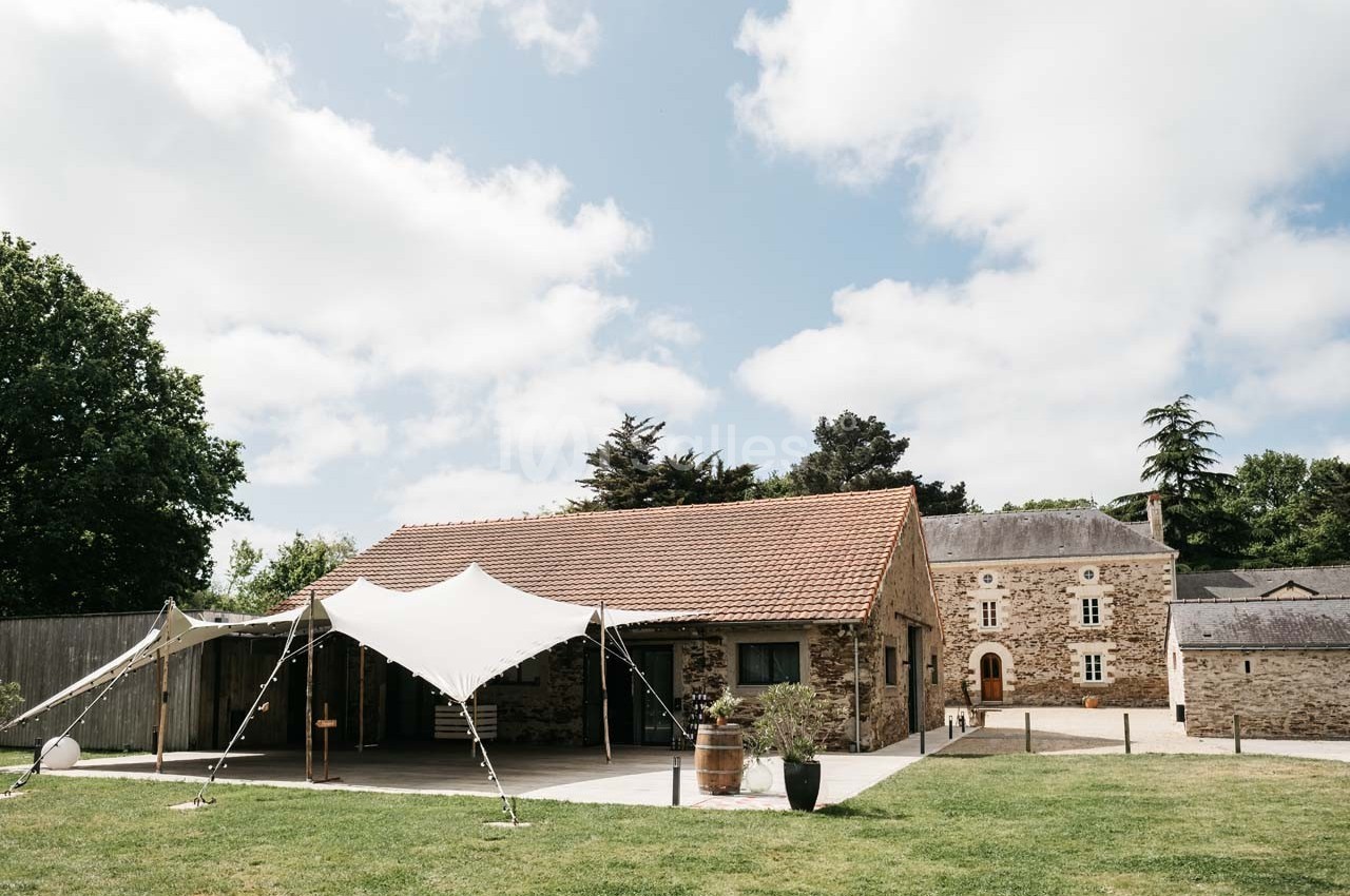 Bâtiment en pierre avec toit en tuiles, tente blanche installée devant, entouré de pelouse et d'arbres.