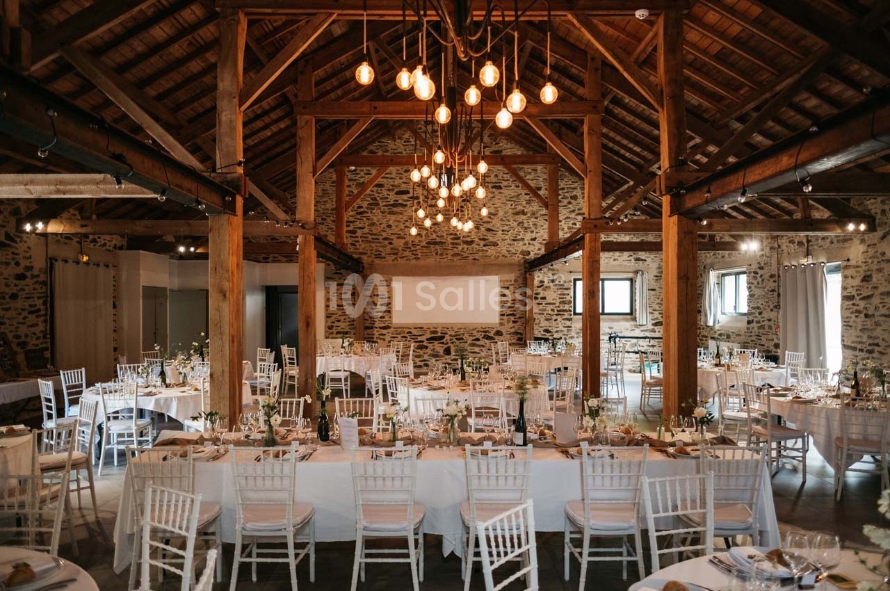 Salle de réception rustique avec poutres apparentes, tables dressées et chaises blanches, éclairée par des suspensions.